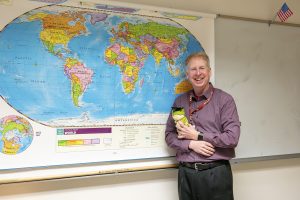 Algonquin history teacher, John Barry poses in front of a world map in the back of his classroom with his stuffed frog, Professor Leland. Professor Leland is Barry's favorite object or stuffed animal on his desk.